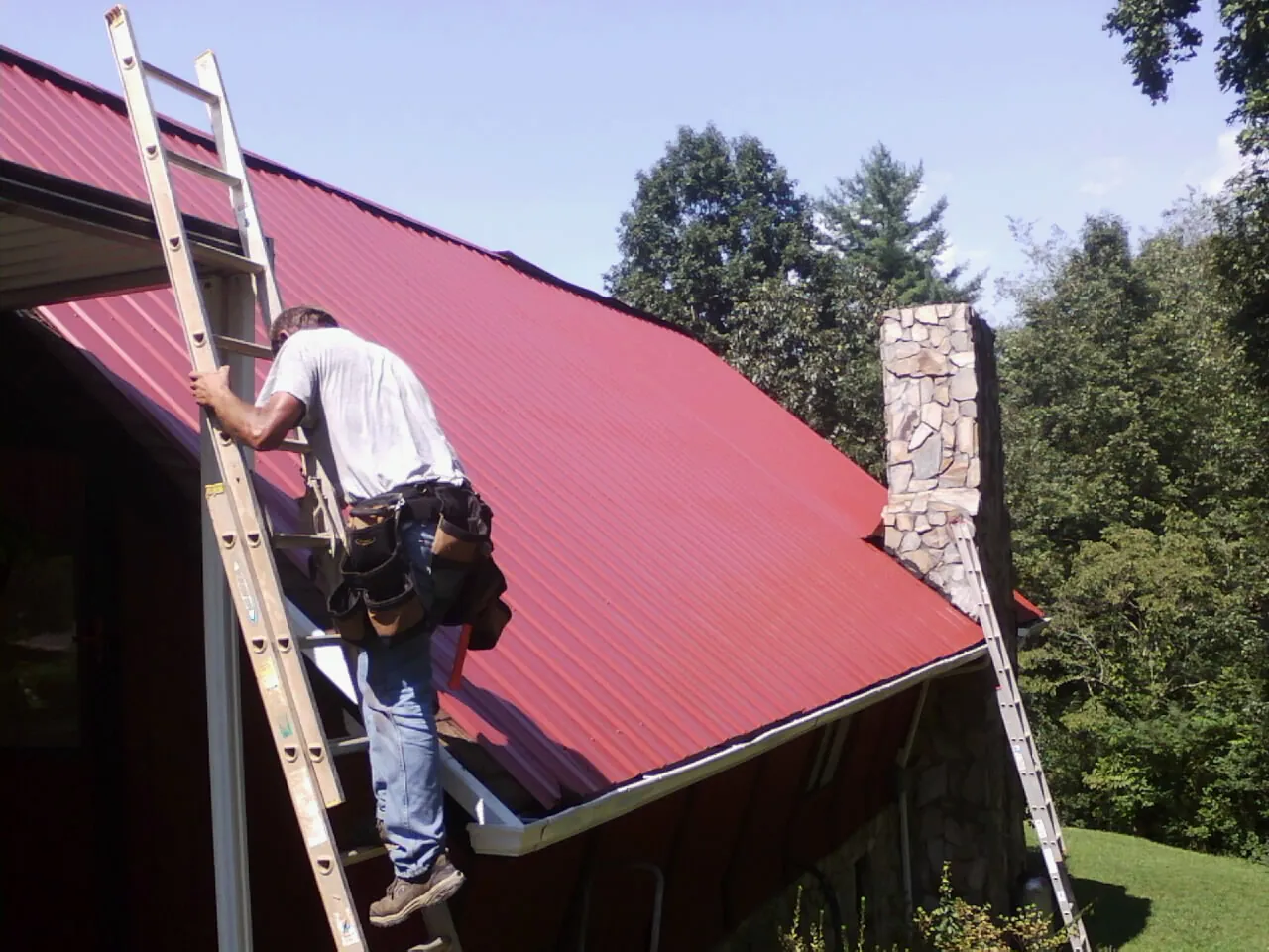 Roofer on a ladder beside a red metal roof and stone chimney — Soffit & Fascia Repair by Roofing Brothers in Auburn